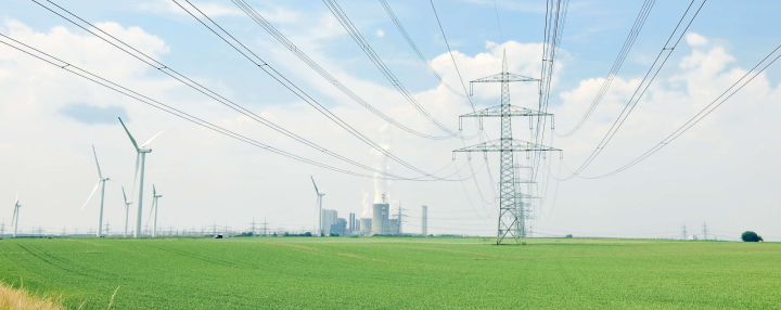 Das Bild zeigt eine Stromtrasse und Windräder in einem grünen Feld - im Hintergrund ein Kraftwerk